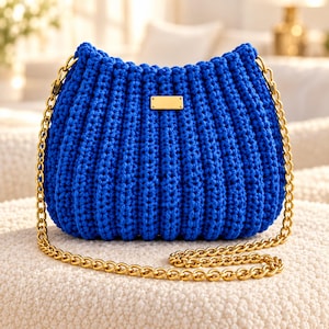 May include: A vibrant royal blue crochet handbag featuring a textured ribbed pattern and a rectangular gold-toned metal plaque on the front. The bag is equipped with a long, polished gold-toned metal chain strap that attaches to the sides.