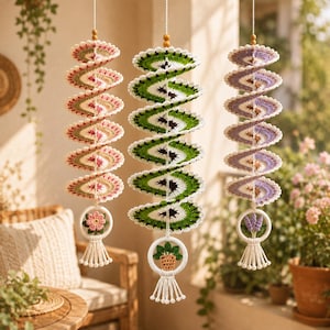 May include: Three hanging spiral crochet decorations suspended by strings. The left features pink and green tones with a flower charm, the centre is green and white with a potted plant charm, and the right is purple and white with a lavender charm.
