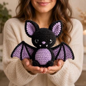 Könnte beinhalten: Ein gehäkeltes Fledermaus-Stofftier mit schwarzem Körper und hellviolettem Bauch, Innenohren und Flügelmembranen. Die Fledermaus hat große schwarze Augen, eine kleine hellviolette Nase und zwei kleine weiße Fangzähne im Mund.
