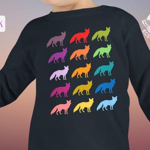 Pode incluir: Uma camisola preta de manga comprida com uma grelha de quinze silhuetas de raposas coloridas em vários tons, incluindo vermelho, amarelo, azul, verde e roxo. O canto superior direito apresenta um logótipo com uma traça e o texto The Ecstatic Moon.
