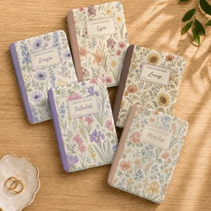 May include: Five rectangular composition books with floral patterns and solid-coloured spines. Each book features a central label with the text "Composition Book" and a name: "Daisie," "Effie," "Lacey," "Tallulah," or "Marley." A small white dish with two gold rings sits nearby.