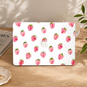 Puede incluir: Una funda para portátil blanca con un patrón repetitivo de acuarelas de fresas rosas con hojas verdes y pequeñas semillas negras. El diseño está impreso en la superficie, con un recorte central para el logotipo de Apple en la tapa del portátil.