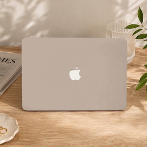 Puede incluir: Un ordenador portátil de color gris claro cerrado con un acabado mate suave y un logotipo de Apple blanco centrado en la tapa. El dispositivo presenta esquinas redondeadas y un diseño rectangular elegante, con una apariencia uniforme.