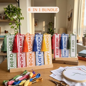 Op de afbeelding: Een 8-in-1 bundel kruissteek-bladwijzers met de teksten READ MORE, ADVENTURES AWAIT, BOOKWORM, LOST IN A BOOK, HAPPY READING, KEEP GOING, STAY STRONG en BE KIND, elk voorzien van een bijpassend gekleurd kwastje en houten standaard.