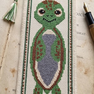 May include: A rectangular bookmark featuring a cross-stitch design of a green turtle with brown spots and a light purple belly. The bookmark is bordered by a green and brown geometric pattern and includes a green and white braided tassel at the top.
