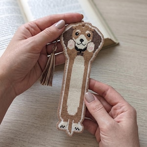 Puede incluir: Un marcapáginas de punto de cruz con forma de perro de cuerpo alargado, pelaje marrón y blanco, collar negro y una pequeña nariz negra. El marcapáginas tiene una borla de color marrón claro en la parte superior y un patrón bordado detallado.
