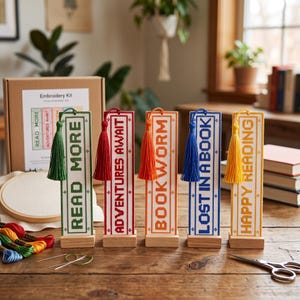 May include: Five rectangular embroidered bookmarks with tassels, each featuring a unique color and text: green READ MORE, red ADVENTURES AWAIT, orange BOOK WORM, blue LOST IN A BOOK, and yellow HAPPY READING, all standing on small wooden bases.