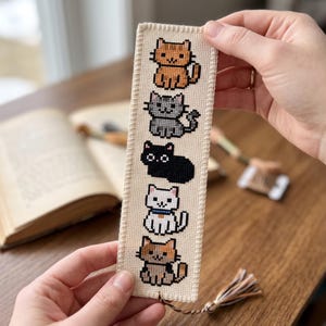 Op de afbeelding: Een rechthoekige crèmekleurige stoffen boekenlegger met een verticaal kruissteekpatroon van vijf verschillende katten op een rij. De katten zijn oranje, grijs, zwart, wit met een blauwe halsband en lapjeskat. Onderaan is een kwastje bevestigd.