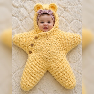 May include: A bright yellow, star-shaped crochet baby bunting with a hood featuring small bear ears. The garment is secured with three brown wooden buttons along the side. A soft, light pink headband with a small bow is worn around the head.