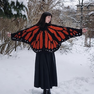 May include: A black and orange crochet cardigan featuring a monarch butterfly wing pattern with white accents along the edges. The garment has long, wide sleeves, a button-down front, and a high neckline, worn over a long black skirt and boots.
