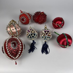 Puede incluir: Una colección de ocho adornos decorativos con diversas formas y texturas. El conjunto incluye esferas de satén rojo con cuentas, dos formas geométricas con borlas azul oscuro y un gran adorno rojo decorado con perlas.