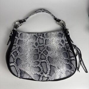 May include: A hobo-style handbag featuring a grey and black snakeskin pattern on the front panel. The bag includes black side panels, a single top handle attached with silver-toned metal rings, and decorative black leather tassels on the side.