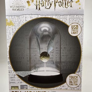 May include: A Harry Potter Wizarding World bell jar light featuring a golden snitch with silver wings inside a glass dome on a black base. The packaging includes text reading USB POWERED LIGHT, TOUCH ACTIVATED ON/OFF FUNCTION, and BELL JAR LIGHT.