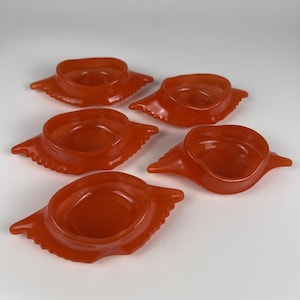 Puede incluir: Un conjunto de cinco cuencos de cristal naranja translúcido con forma de cangrejo. Cada cuenco presenta un recipiente circular central con patas de cangrejo estilizadas que se extienden por los lados. El cristal tiene un acabado liso y brillante.