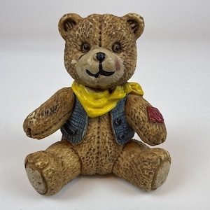 May include: A brown textured teddy bear figurine wearing a blue vest over a yellow neckerchief. The bear has a small red patch on its left arm, dark eyes, and a black stitched nose and mouth. The figure is seated in an upright position.