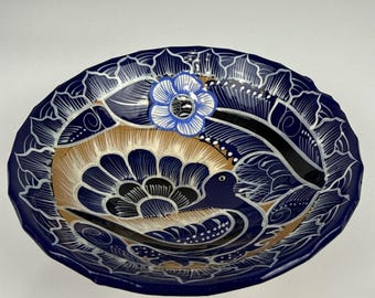 Puede incluir: Un cuenco de cerámica redondo con un borde azul profundo y contornos florales blancos. El centro muestra un motivo de ave pintado a mano en blanco y negro sobre un fondo beige, acentuado por una flor azul y blanca en el borde superior.