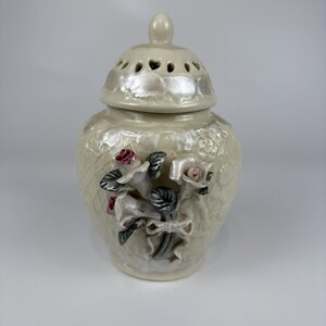 May include: A cream-colored ceramic jar with a matching lid featuring a perforated top. The jar has a textured lattice pattern and is decorated with three-dimensional sculpted roses in shades of pink and grey, accented with a ribbon detail.