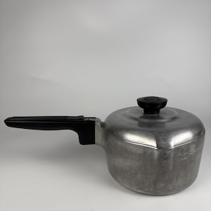May include: A silver-coloured aluminium saucepan with a matte finish, featuring a long black plastic handle and a matching black plastic knob on the circular lid. The pot has a rounded body and a flat base, designed for stovetop cooking use.