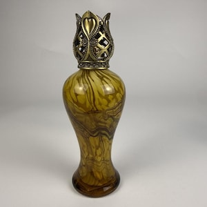 May include: A glass fragrance lamp with a swirling amber and brown marbled pattern. The top features an ornate, metallic gold-toned crown-shaped cap with a lattice design and heart-shaped accents, sitting atop a slender, curved glass body.