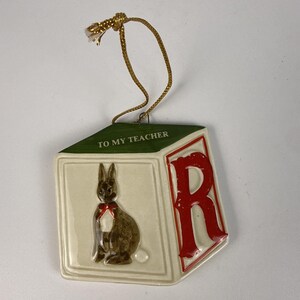May include: A ceramic block-shaped ornament featuring a green top with the text TO MY TEACHER, a brown rabbit illustration with a red bow, and a large red letter R on the side. The ornament hangs from a gold-colored cord.