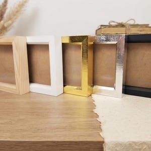 May include: A set of five square shadow box frames arranged in a row. The frames feature different finishes including natural light wood, matte white, reflective gold, reflective silver, and matte black. Each frame has a deep profile and brown backing.