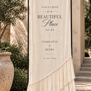 May include: A tall, cream-colored fabric banner suspended on a black metal frame. The banner features black printed text: A NEW CHAPTER BEGINS, TOGETHER is a BEAUTIFUL Place TO BE, CHARLOTTE & HENRY, and 22.06.2026. The fabric has a diagonal ruffle detail.