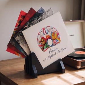 May include: A black wooden vinyl record display stand holding a collection of records, with the Queen A Night at the Opera album cover featuring a colorful crest and text prominently displayed in the front slot of the rack.