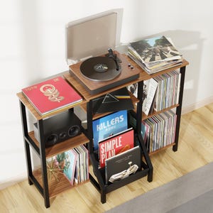May include: A multi-tiered wooden record player stand with a black metal frame. The unit features shelves for vinyl records, a speaker, and a turntable on top. Visible album covers include Toto, Pink Floyd, The Killers, and Simple Plan.