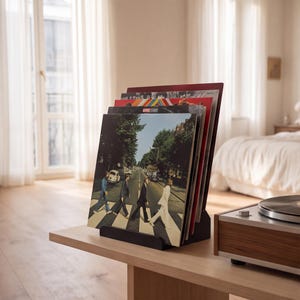 May include: A black vinyl record storage stand holding a collection of music albums. The front-most record cover features a photograph of four people walking across a street crosswalk. The stand is constructed with a solid base and angled back.