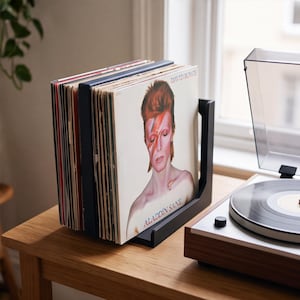 May include: A black metal vinyl record storage rack holding a collection of records, with the David Bowie Aladdin Sane album cover prominently displayed at the front. The rack sits next to a turntable on a wooden surface.