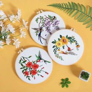 Può includere: Tre telai da ricamo circolari in legno con tessuto bianco e disegni ricamati dettagliati. Ogni telaio mostra un colibrì colorato che interagisce con varie composizioni floreali, tra cui glicini viola, fiori gialli e fiori rossi.