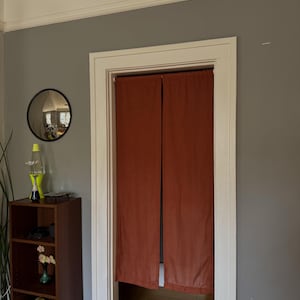 Pode incluir: Um par de cortinas de porta de tecido na cor ferrugem sólida penduradas dentro de uma moldura de porta branca. As cortinas são retangulares, opacas e apresentam um design simples e reto que cobre a abertura da porta de cima a baixo.