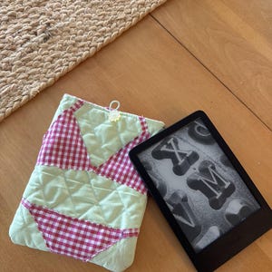 May include: A quilted fabric sleeve featuring a light green background with a red and white gingham bikini pattern appliqué, including a small top loop. Beside it sits a black e-reader displaying a screen with the letters X and M in a bold font.