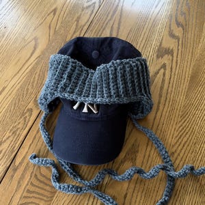 May include: A dark navy blue baseball cap featuring a white embroidered NY logo on the front, adorned with a thick, dark gray crocheted ear warmer band that wraps around the crown and includes long, dangling crocheted ties on each side.