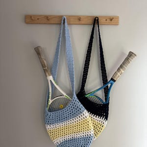 May include: Two crochet bags hanging on a wooden rack, each containing a tennis racket. One bag features horizontal stripes of light blue, white, and yellow. The other bag is solid dark navy blue with a white and yellow striped base section.