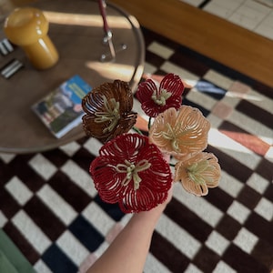 May include: A bouquet of five handcrafted flowers made from small glass beads. The flowers feature petals in shades of deep red, dark brown, and light peach, with pale green beaded centres and stems, arranged in a cluster.