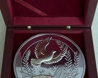 May include: A circular silver-toned commemorative coin featuring an embossed dove in flight surrounded by laurel branches. A banner at the bottom displays the text PEACE 2003. The coin is presented inside a dark red velvet-lined wooden box.