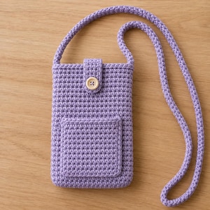Può includere: Una custodia a tracolla per telefono in uncinetto color lavanda, caratterizzata da un corpo rettangolare con tasca frontale, una lunga tracolla intrecciata abbinata e un bottone in legno chiaro su una piccola patta che chiude lo scomparto.