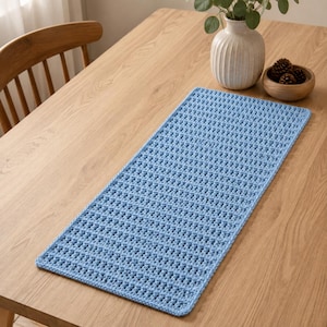 May include: A rectangular light blue table runner featuring a textured crochet stitch pattern. The runner is laid flat on a wooden surface, showcasing a consistent woven design with a repeating grid-like structure throughout the entire piece.
