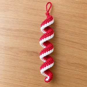 May include: A spiral-shaped crochet ornament featuring alternating red and white yarn stripes. The piece is constructed with a tight, textured stitch pattern and includes a small, integrated loop at the top for hanging purposes.
