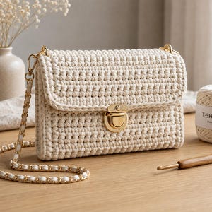May include: A cream-colored crochet handbag featuring a rectangular shape, a fold-over flap, and a gold-tone metal push-lock closure. The bag includes a matching cream crochet and gold chain shoulder strap, with a 6.0mm crochet hook nearby.