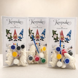 Könnte beinhalten: Drei identische Keepsake Kits Gnome Painting Kit Verpackungen in durchsichtiger Folie. Jedes Set enthält drei unbemalte weiße Keramik-Gnomfiguren, eine Auswahl kleiner Farbtöpfe in verschiedenen Farben und einen Pinsel mit blauem Stiel.