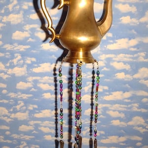 May include: A brass-colored teapot wind chime with a decorative handle and spout. Colorful beaded strands hang from the base, with spoons suspended at the bottom. The background is a blue sky with white clouds.