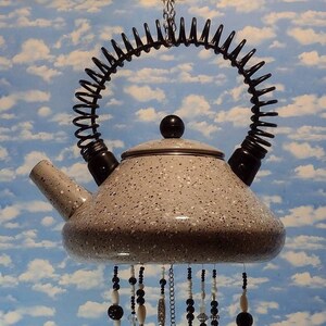 May include: A whimsical wind chime crafted from a speckled gray teapot, adorned with black and white beads, and hanging silverware. The teapot has a black handle and a spring-like design. Silverware includes forks, spoons, and a bell.