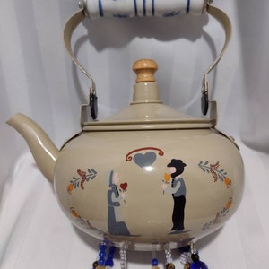 May include: Tan enamel teapot with a white and blue floral handle. The teapot features a painted scene of two figures holding flowers, with a heart above them. Decorative blue and clear beads hang from the bottom.