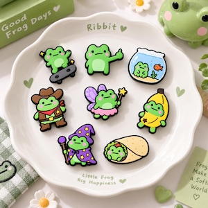 May include: A set of eight colourful frog-themed shoe charms arranged on a white scalloped plate. The designs include a frog on a skateboard, a frog in a fishbowl, a cowboy frog, a fairy frog, a banana frog, a wizard frog, and a frog in a burrito.
