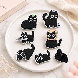 May include: A collection of seven black cat pins featuring various designs: one with a green sprout, one with a small purple bug, one holding a silver knife, one wearing a slice of bread, and three in different playful poses with wide white eyes.