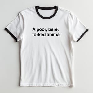 以下が含まれることがあります： 襟と袖口に黒いトリムがあしらわれた白いリンガーTシャツ。胸の中央には「A poor, bare, forked animal」という黒いテキストがプリントされています。半袖で、標準的なクルーネックのデザインです。