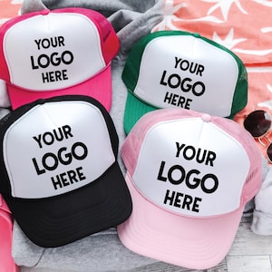 May include: Four trucker hats in pink, green, black, and light pink, each featuring a white front panel with the black text YOUR LOGO HERE. These caps have mesh back panels and solid brims, designed for custom branding and promotional use.