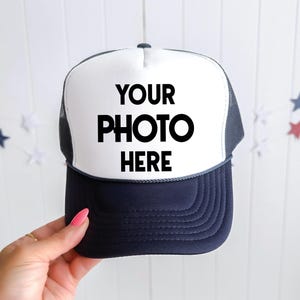 May include: A navy blue and white trucker hat featuring a foam front panel with the black text YOUR PHOTO HERE printed in the center. The cap includes a dark navy blue brim and a mesh back panel, designed with a structured crown and adjustable strap.